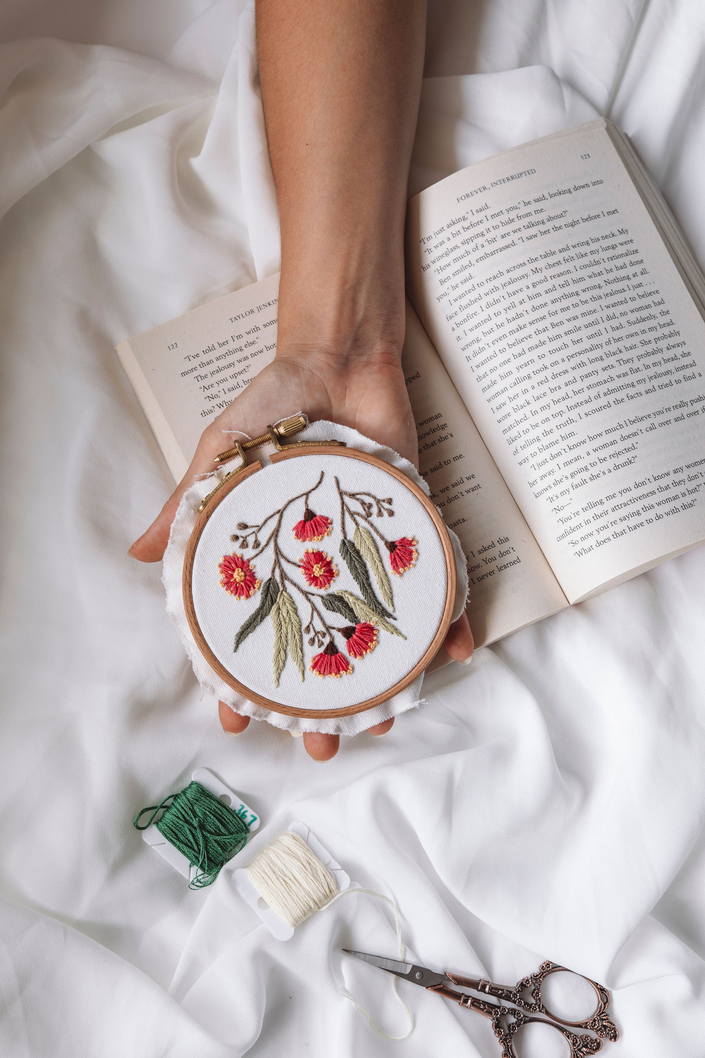 Person holding an embroidery hoop stitched with red flowers and green leaves over an open book on white fabric