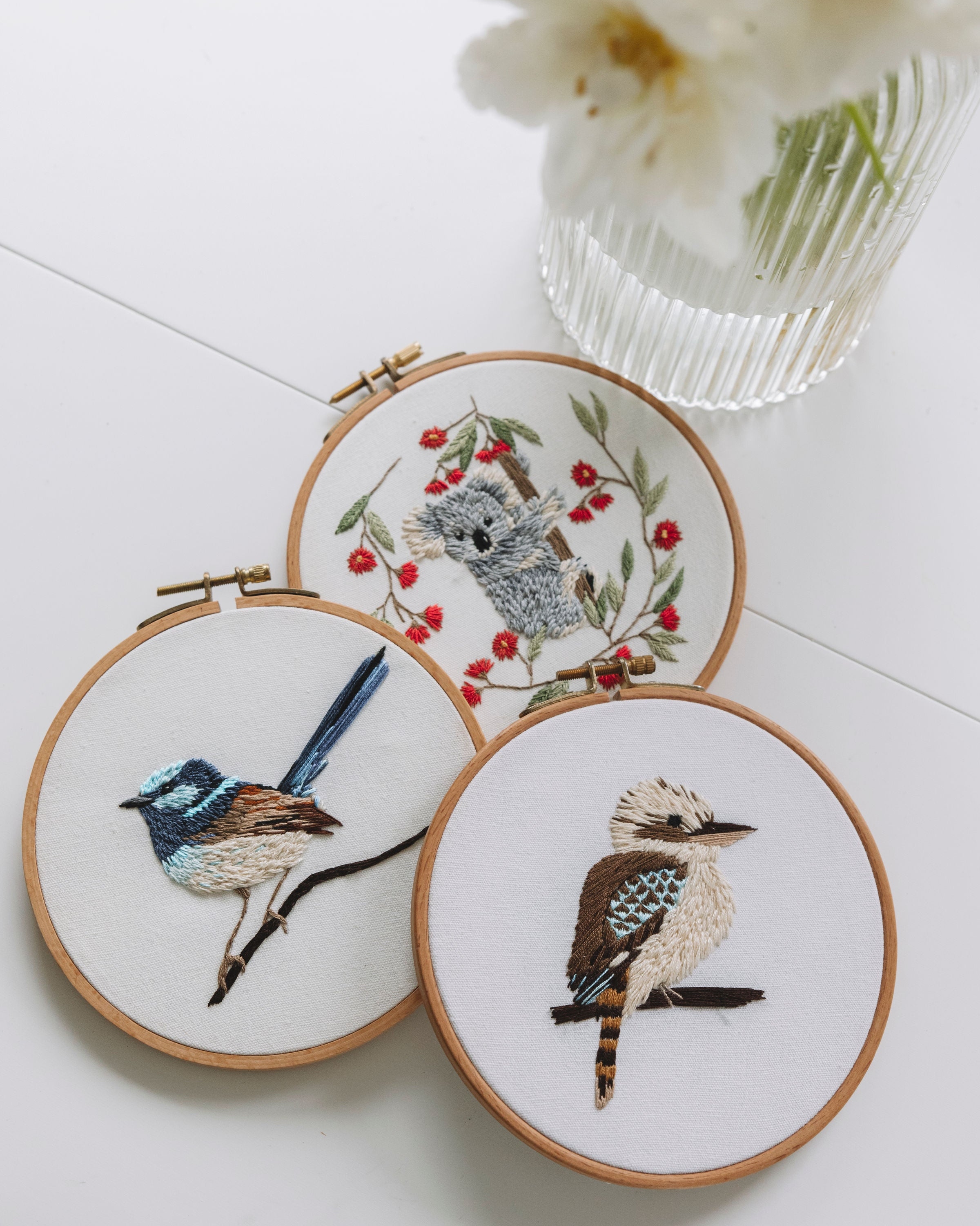 Three wooden embroidery hoops with detailed bird and koala designs arranged near a clear textured vase with white flowers