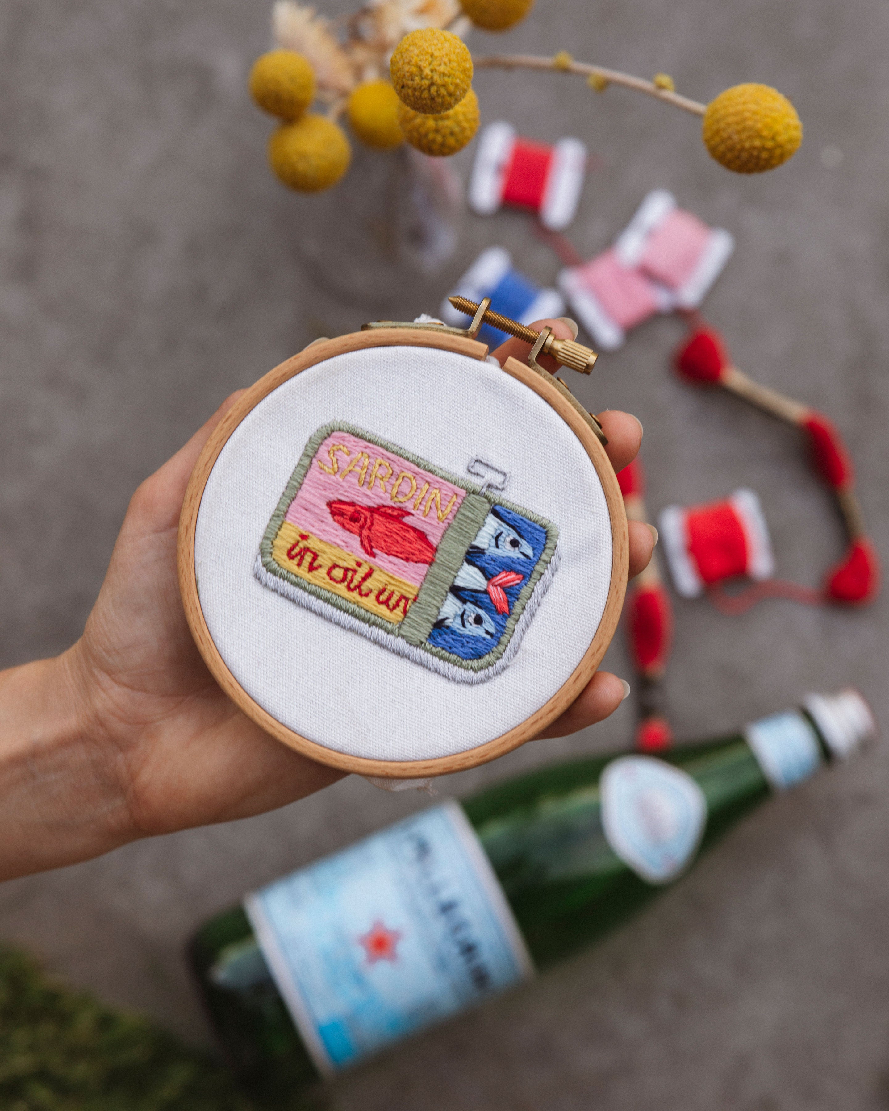 Mini Sardines Tin embroidered patch held in hand with colorful threads and bottle in the background.