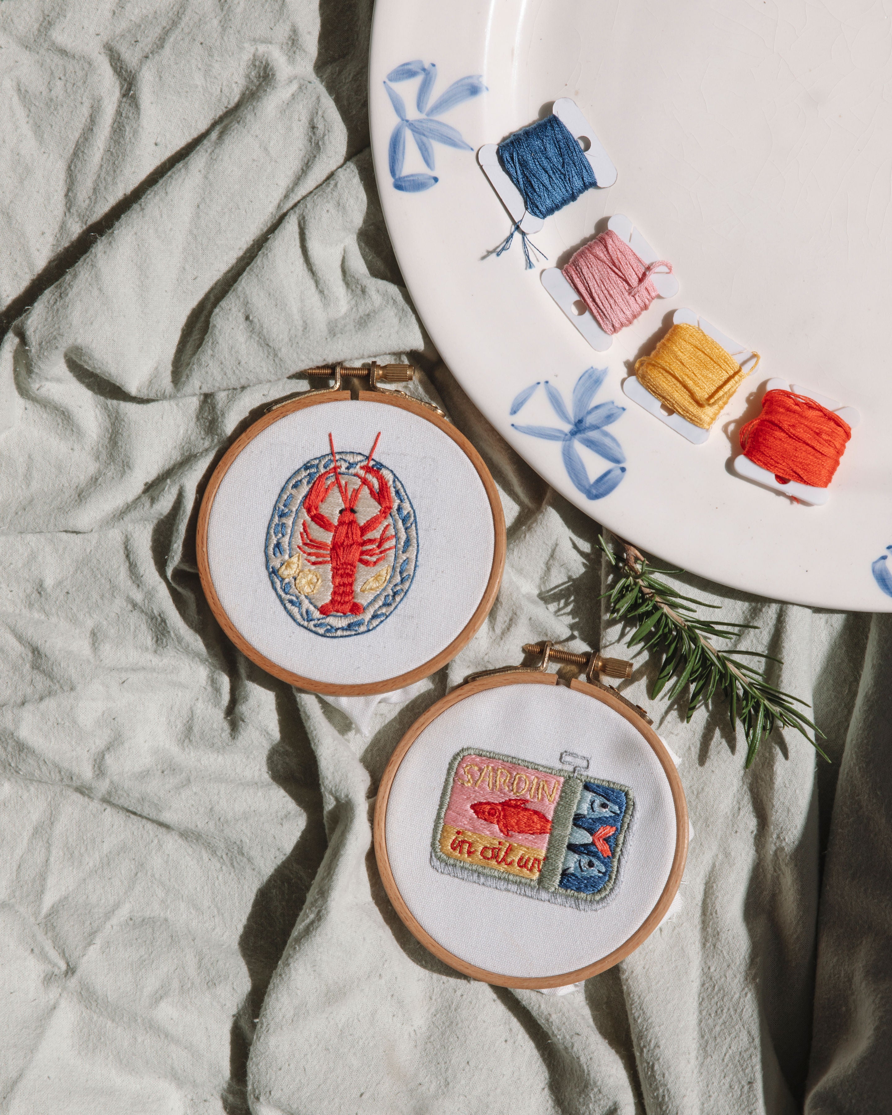 Mini Sardines Tin embroidery artwork displayed in wooden hoops with colorful thread and rosemary on fabric background