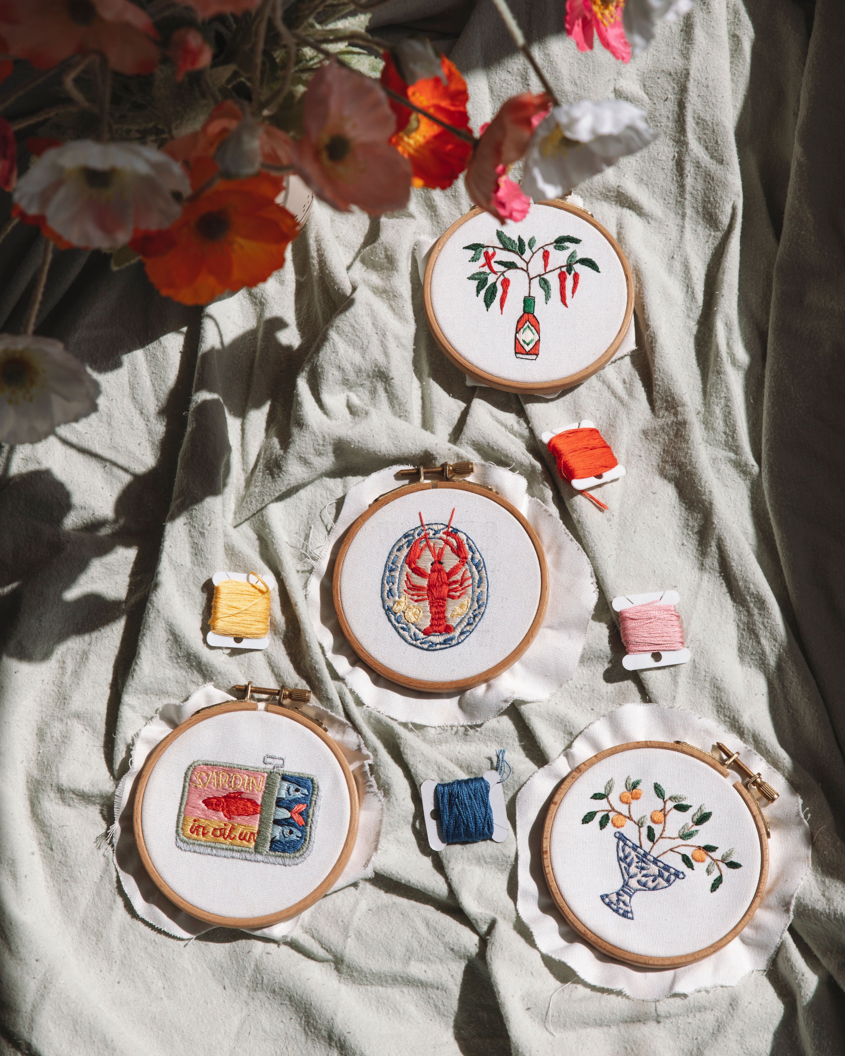 Mini Sardines Tin embroidery design in a wooden hoop surrounded by colorful thread and flowers on fabric background