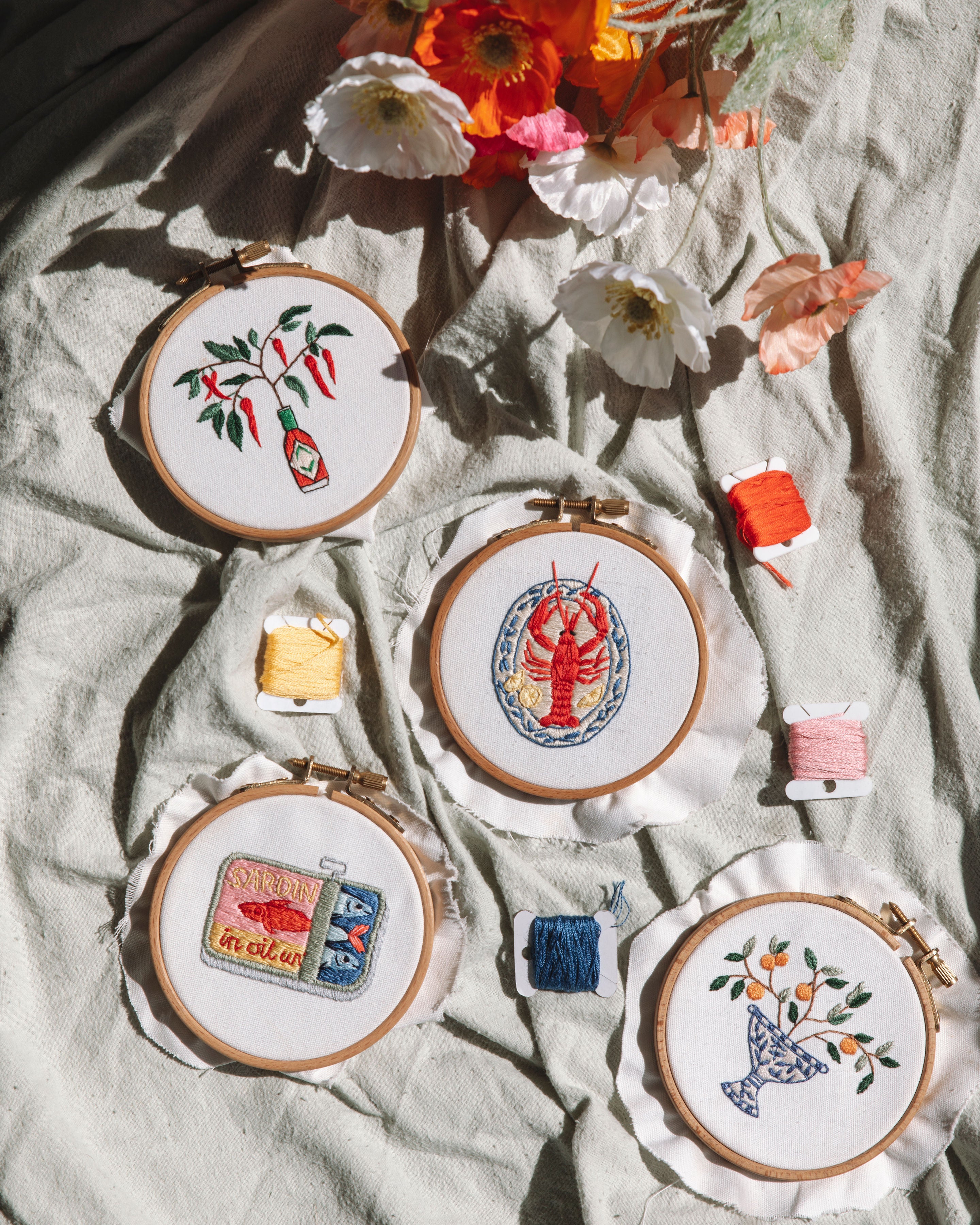 Embroidery hoops featuring Italian-themed designs with hot sauce, lobster, sardines and citrus patterns with colorful thread spools