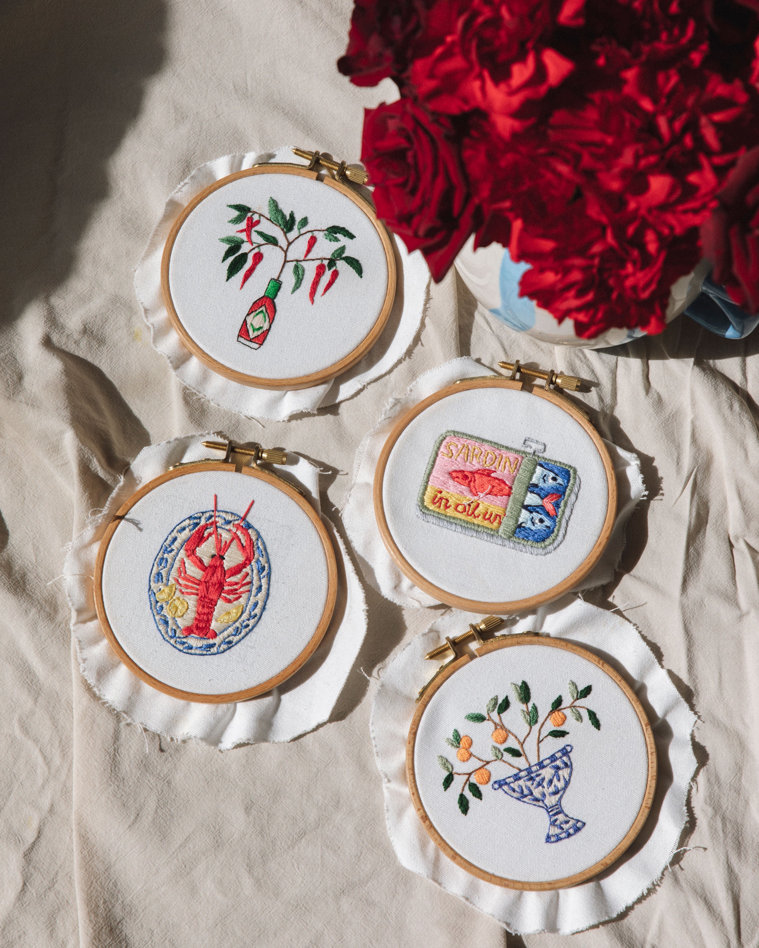 Mini Sardines Tin embroidered artwork in hoop next to floral and lobster designs on fabric with red flowers nearby