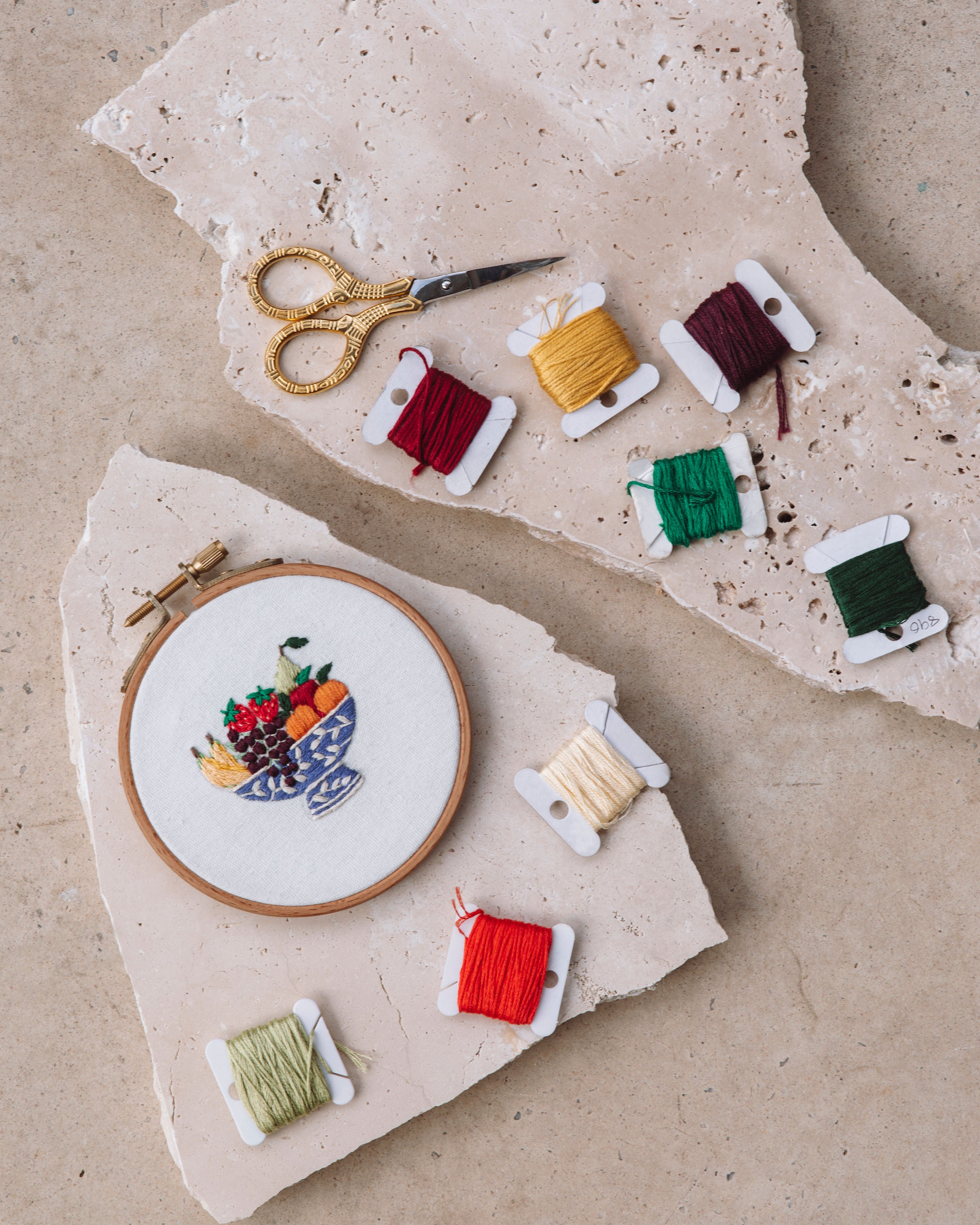Girls Stitch Night Bundle showing embroidered fruit bowl in wooden hoop with colorful threads and vintage scissors on stone slabs