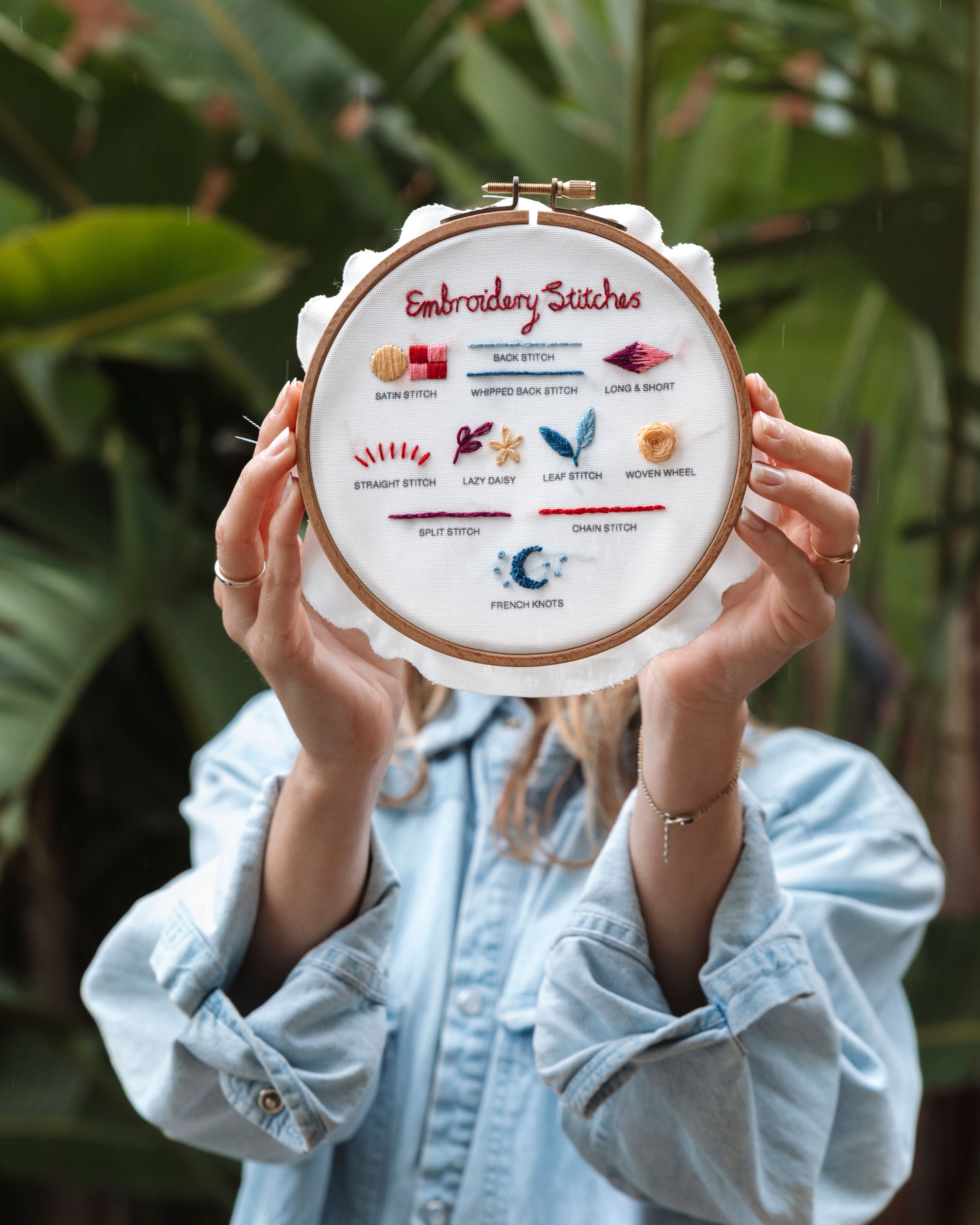 Person holding Beginner Embroidery Bundle showing colorful labeled embroidery stitch examples in wooden hoop outdoors