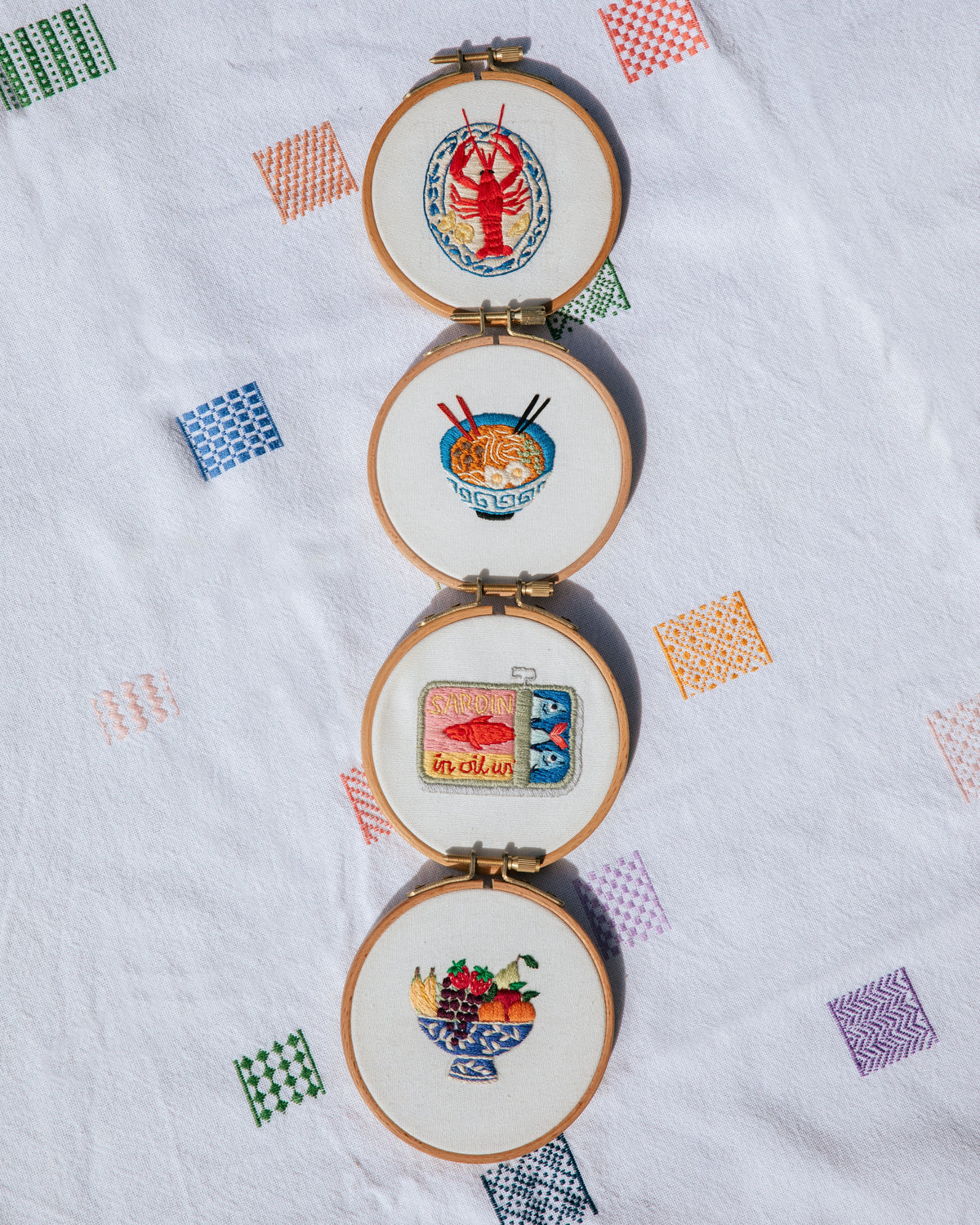 Four embroidery hoops with colorful stitched designs including lobster, ramen bowl, sardines tin, and fruit bowl on fabric background