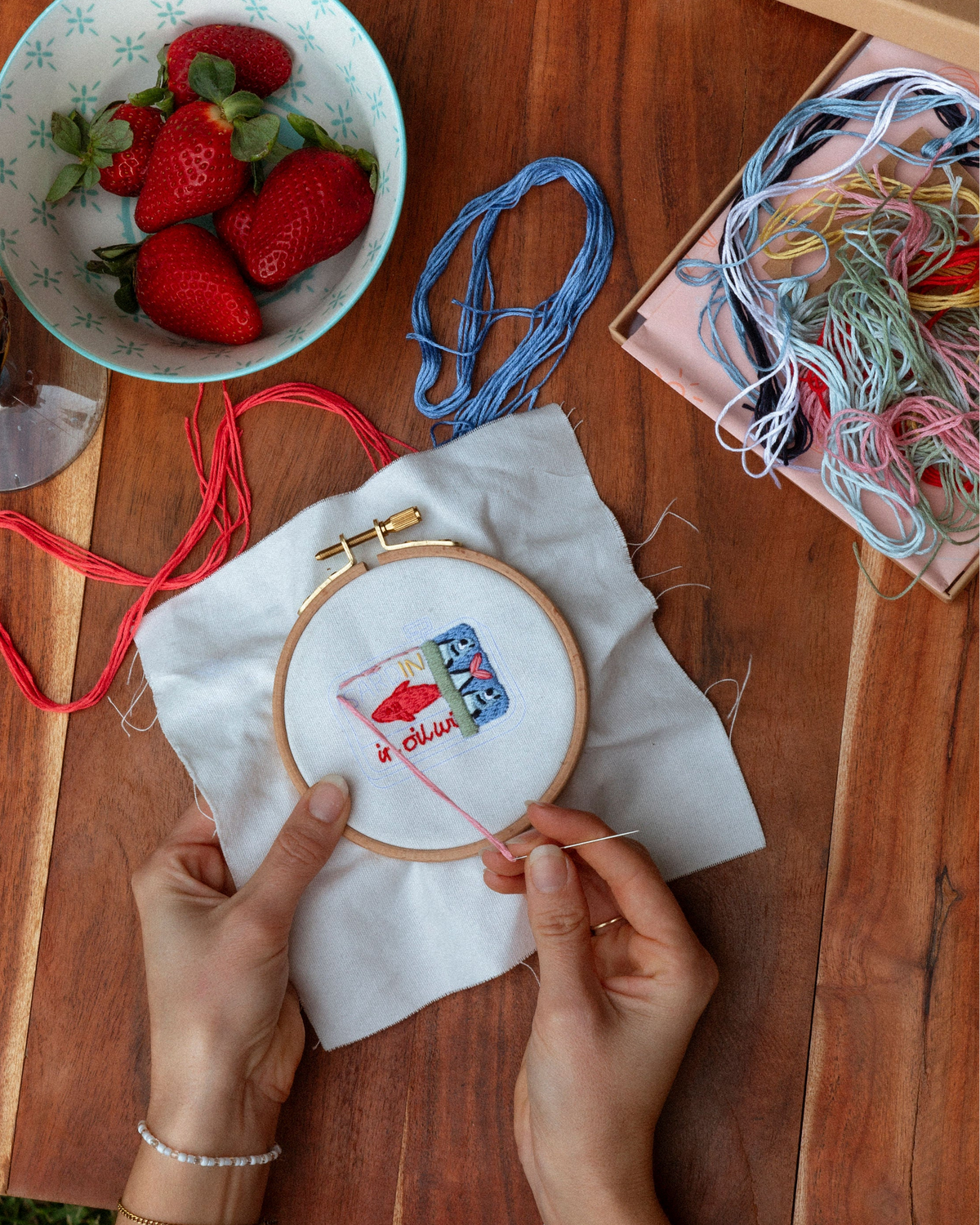 Hands stitching a detailed small embroidery design on fabric with colorful threads and fresh strawberries nearby on wood surface