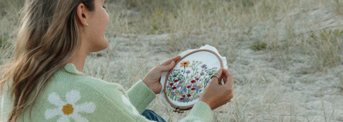 Person wearing green sweater with daisy pattern holding wooden embroidery hoop with colorful floral design outdoors on sandy grass