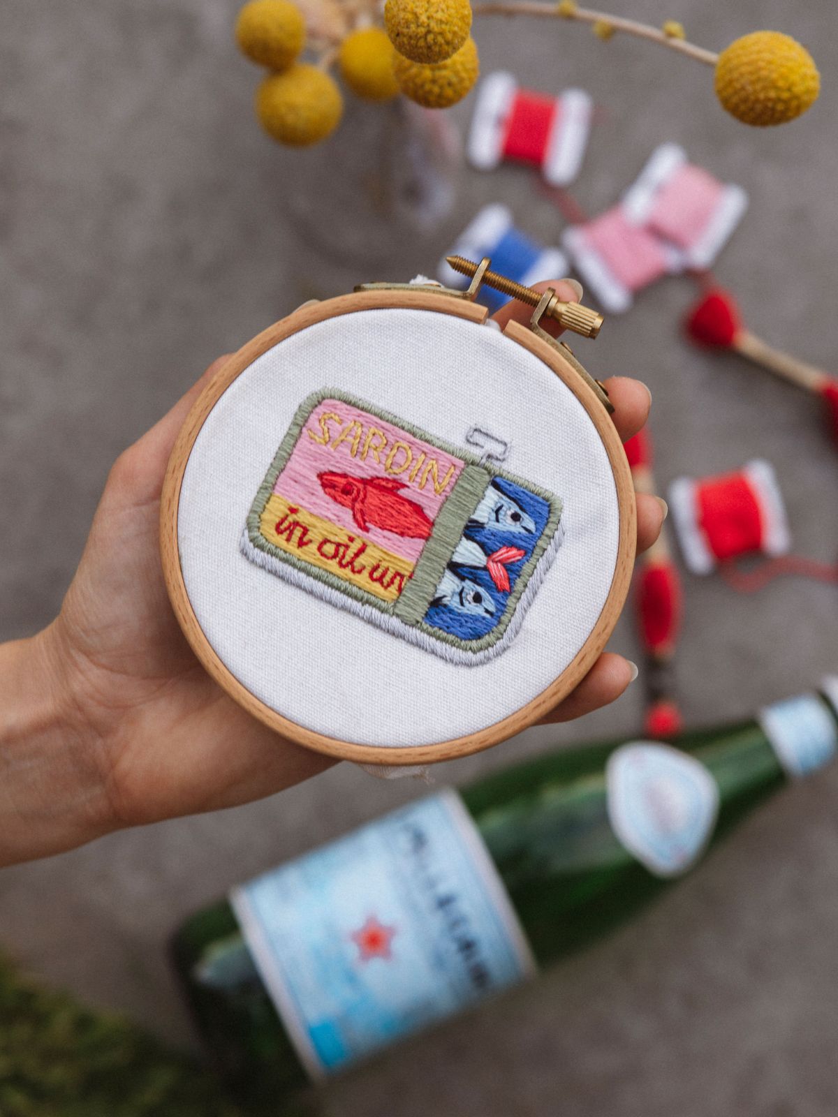 Hand holding wooden embroidery hoop with colorful sardine tin design stitched on white fabric with thread spools nearby