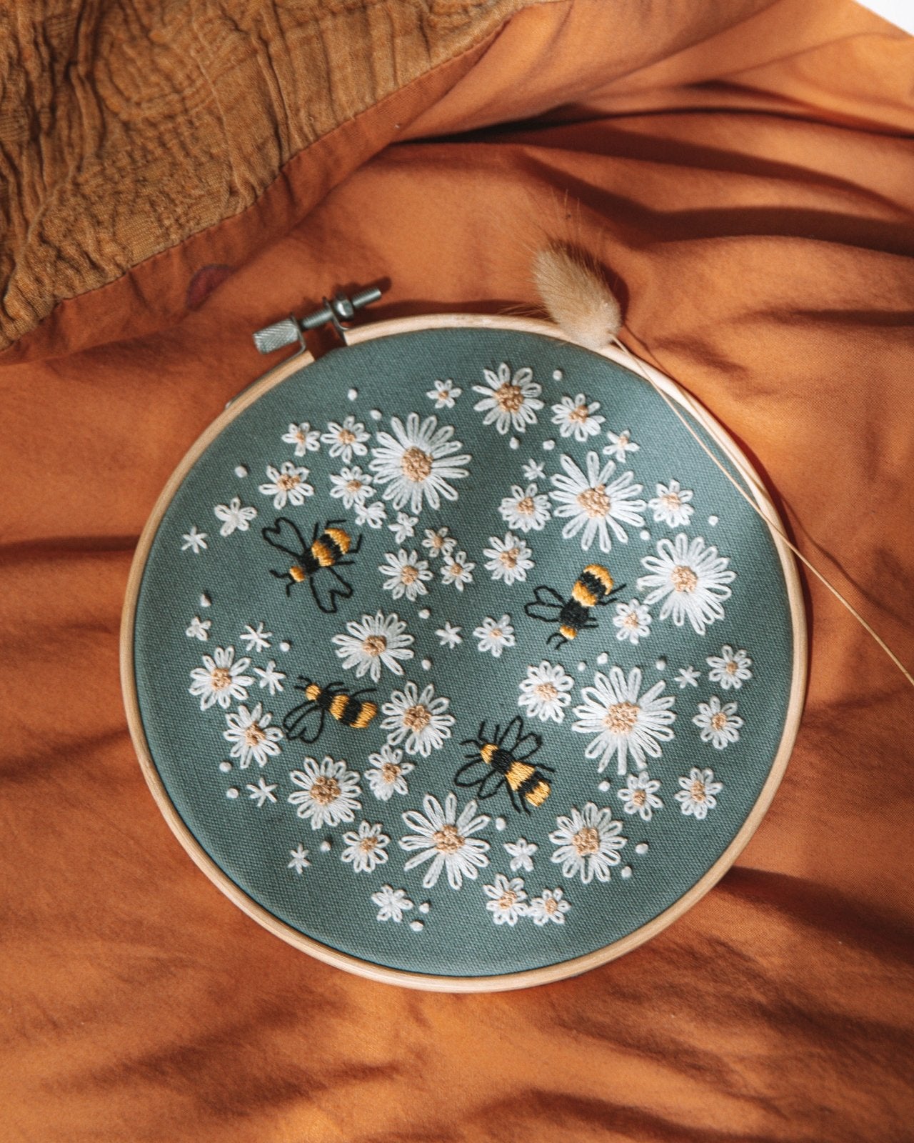 Completed Busy Bumble Bee embBusy Bumble kit on an orange bed sheet. Design is on dark Blue-Grey fabric and has white flowers all over it and 4 bees in amongst them.