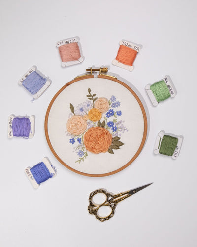 This is a flat-lay of a completed floral embroidery piece with 7 embroidery floss and scissors spaced around it in a circle 