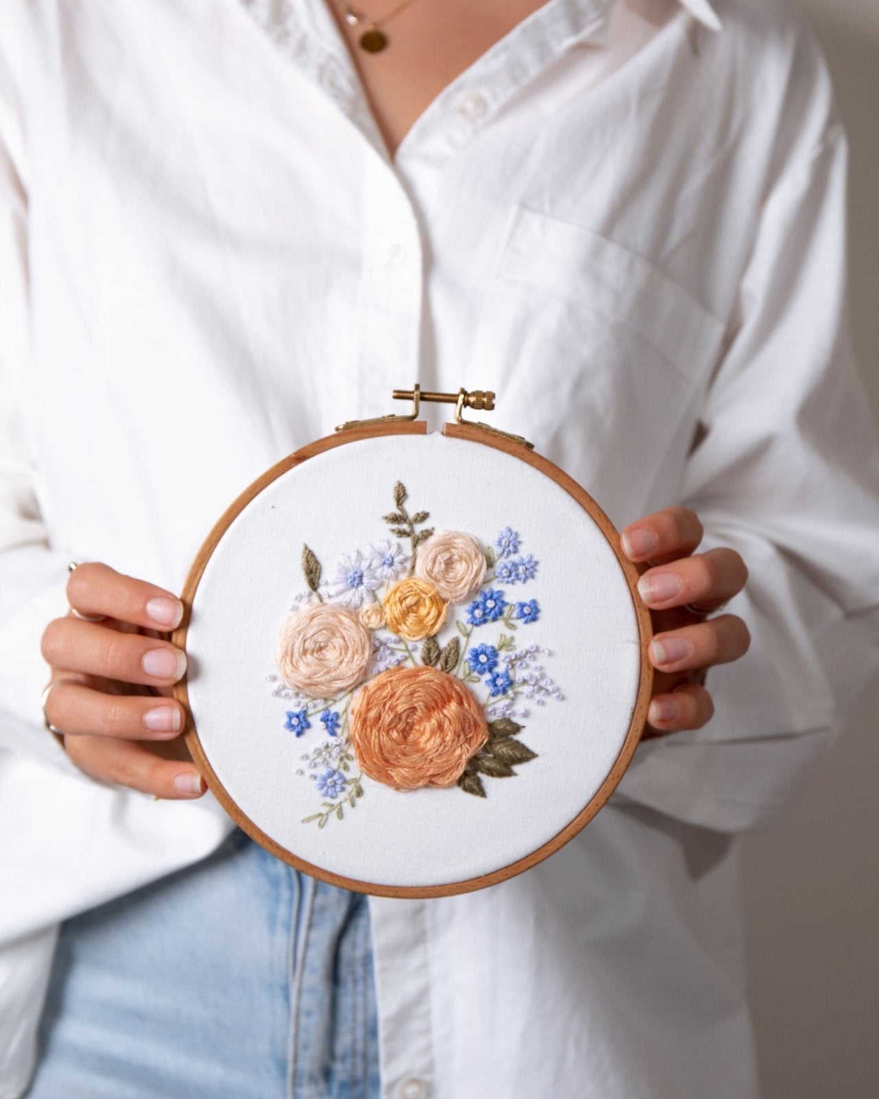 Girl in white shirt and light blue jeans holding completed floral embroidery in a wooden embroidery hoop. The embroidery has pink and blue flowers with a lot of texture in the roses. 