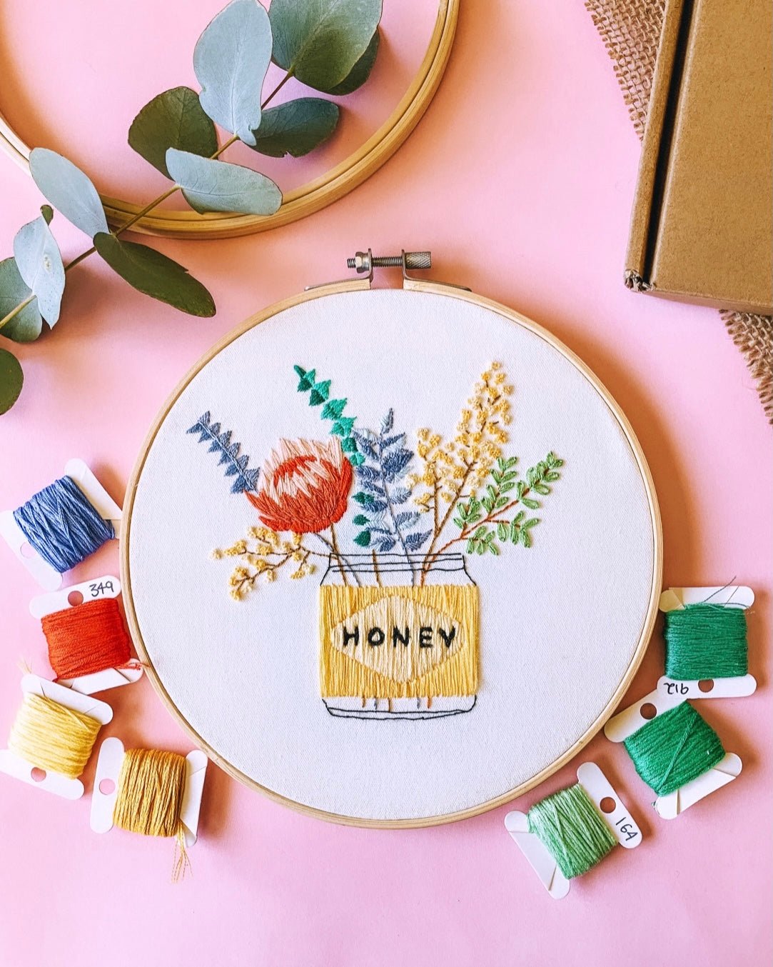 Finished embroidery piece of yellow honey jar with australian natives sitting in it including wattle and gum leaves, sitting on a pink background